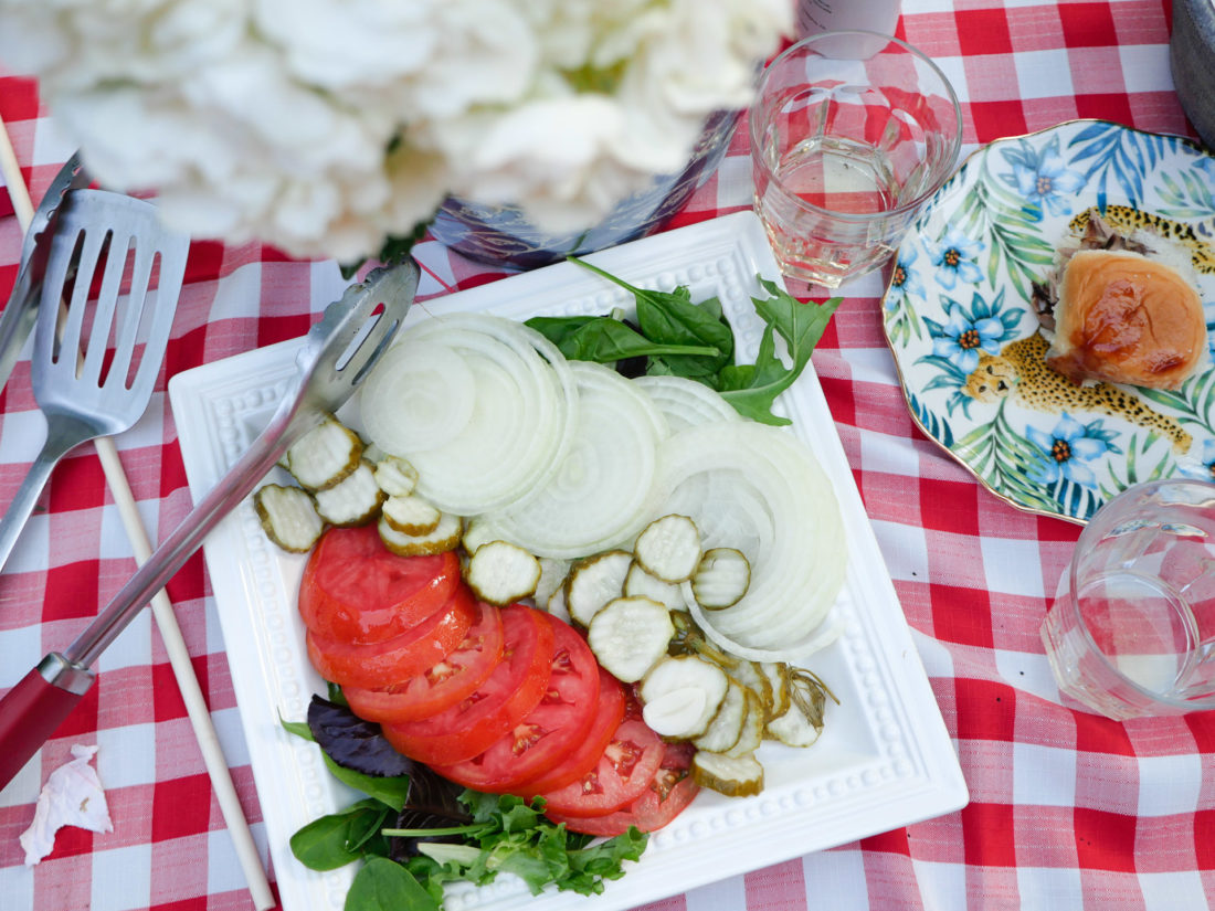 Delicious food and hamburger fixings sit on a red and white checked tablecloth at a garden party on the fourth of july