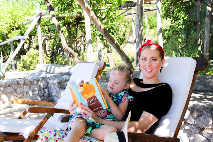 Eva Amurri Martino sits with daughter Marlowe on a chair in Amalfi reading her newest HEA Book Club pick: Eleanor Oliphant Is Completely Fine
